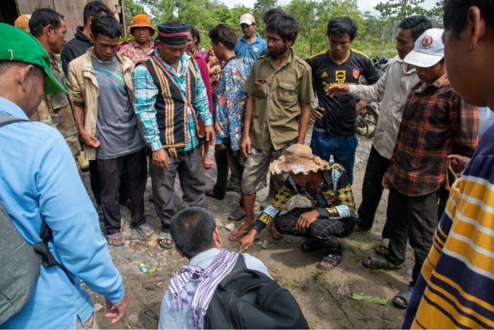 ពេលទៅដល់ទីសក្ការៈក្នុងដីសម្បទានសង្គមកិច្ច លោក គីង វីរៈសិទ្ធ ទីប្រឹក្សាជាន់ខ្ពស់ផ្នែកគ្រប់គ្រងហានិភ័យបរិស្ថានជាតិ ជាអ្នកតំណាងគម្រោង LASED III (ខាងមុខ) បានចាប់ផ្តើមគូរព្រំប្រទល់ និងផែនទីលើដី ខណៈដែលប្រធានសហគមន៍ពូនង ឈ្មោះ លីន ម៉ៅ (ពាក់អាវកាក់ និងមួកប្រពៃណី) និងសមាជិកសហគមន៍ផ្សេងទៀតឈរជុំវិញគាត់ ដើម្បីបង្ហាញពីកង្វល់របស់ពួកគេកាលពីថ្ងៃទី១៤ ខែកក្កដា។ (ខេមបូចា/ Andrew Califf)